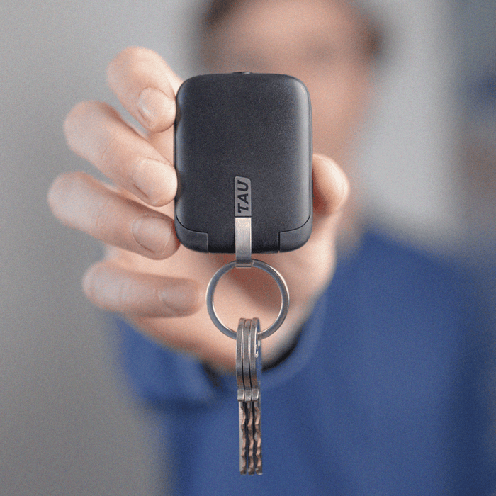 TAU® - Always-charged power bank - TAU® - Always-charged power bank - Charcoal Black - Compact charcoal black power bank with metal keyring attached, held in hand against blurred background. - Rolling Square TAU® - Always-charged power bank - Charcoal Black - Compact charcoal black power bank with metal keyring attached, held in hand against blurred background. - Rolling Square