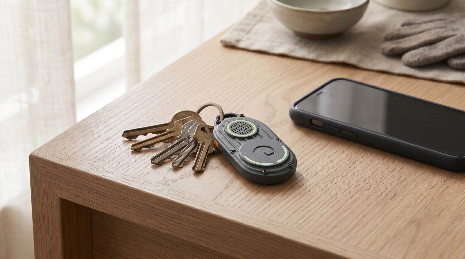 Keyring with house keys and a compact Bluetooth key tracker on an entry table next to a smartphone