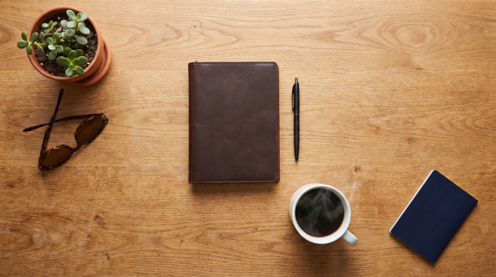 A minimalist flat lay of a leather notebook, coffee, sunglasses, and a passport on a warm wood desk — the kind of calm you get when your tech pouch is not the chaos of the trip